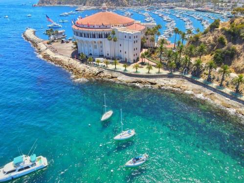 Aerial view of a coastal building with a red dome, surrounded by boats and clear blue water near a marina.