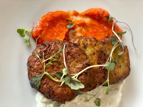 Two golden-brown fritters on a plate with red sauce and a dollop of creamy sauce, garnished with microgreens.