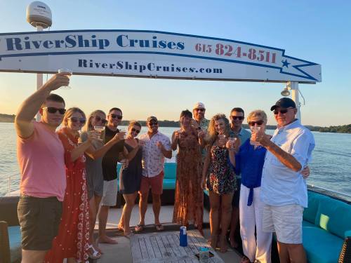 A group of people on a boat, raising drinks and smiling, with a scenic river view in the background.