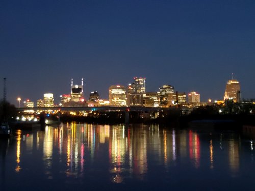 Nashville skyline at night, illuminated buildings reflecting on the calm river below.