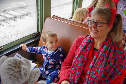 A smiling child in pajamas sits next to a woman in a red sweater and scarf, both enjoying a cozy train ride.