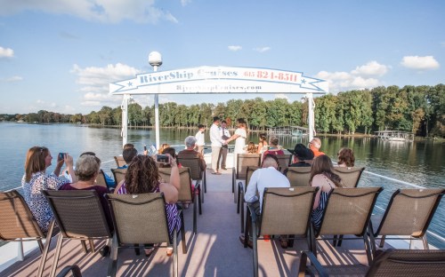 A group of people on a boat enjoying a scenic river view, with trees and a sign for RiverShip Cruises in the background.