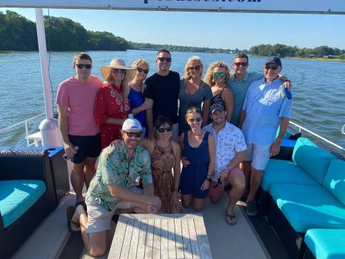 A group of friends poses together on a boat, smiling, with a scenic river view in the background.