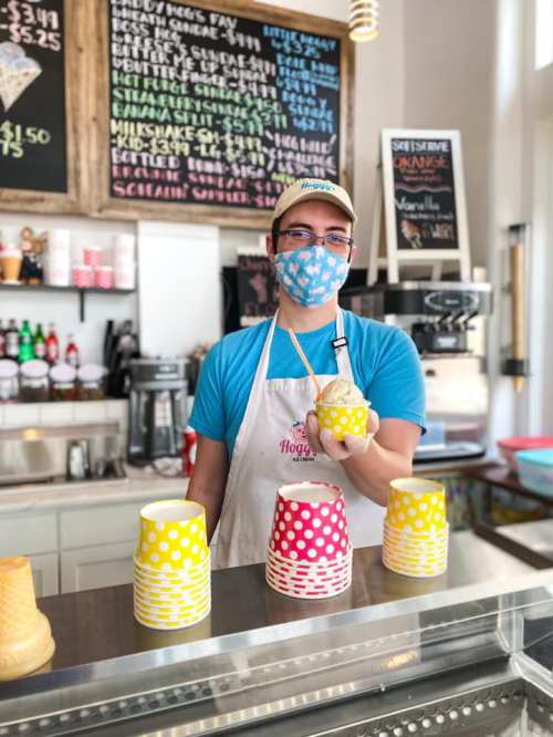A smiling employee in a mask holds a cup in a colorful ice cream shop with polka dot cups and a menu in the background.