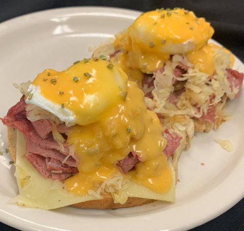 Two open-faced sandwiches topped with corned beef, sauerkraut, melted cheese, and poached eggs, drizzled with cheese sauce.