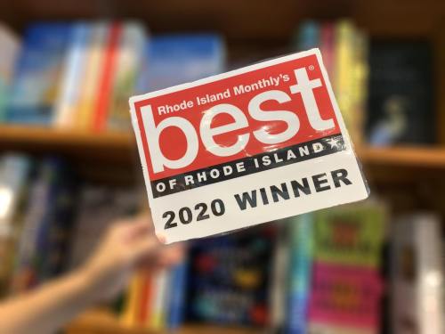 A hand holds a "Best of Rhode Island 2020 Winner" sticker in front of a blurred bookshelf.