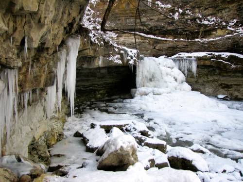 A frozen waterfall surrounded by icy rocks and icicles, set in a rocky, snow-covered landscape.
