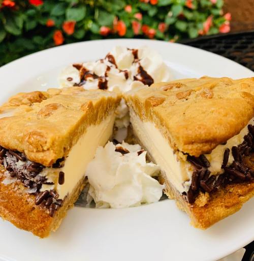 A dessert featuring a cookie sandwich filled with ice cream, topped with whipped cream and chocolate drizzle.