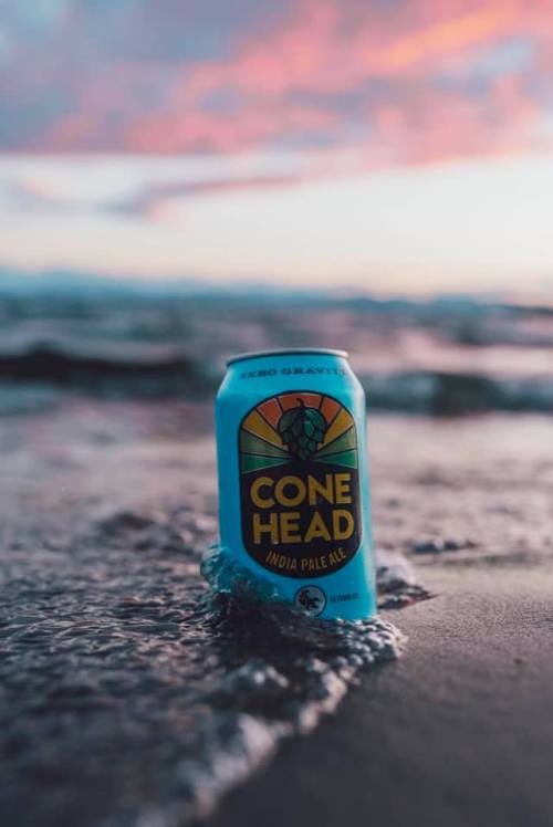 A can of Cone Head India Pale Ale sits on a beach, partially submerged in water with a colorful sunset in the background.