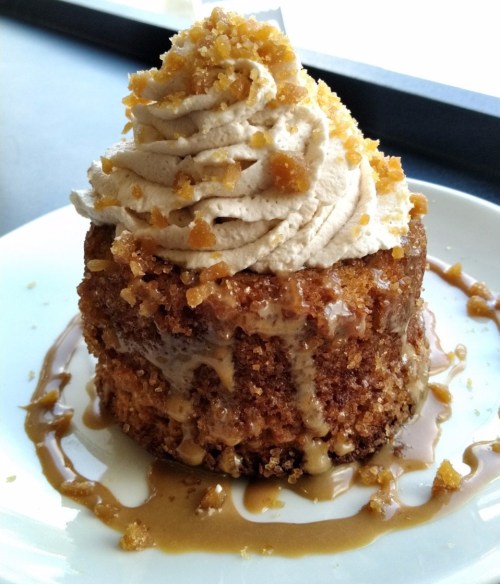 A moist cake topped with whipped cream and crushed toffee, drizzled with caramel sauce on a white plate.