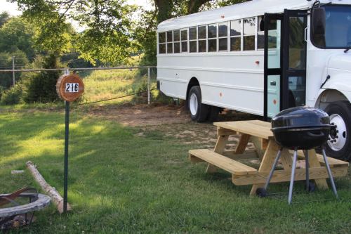 A white bus parked near a picnic table and grill, with a sign reading "216" in a grassy area surrounded by trees.