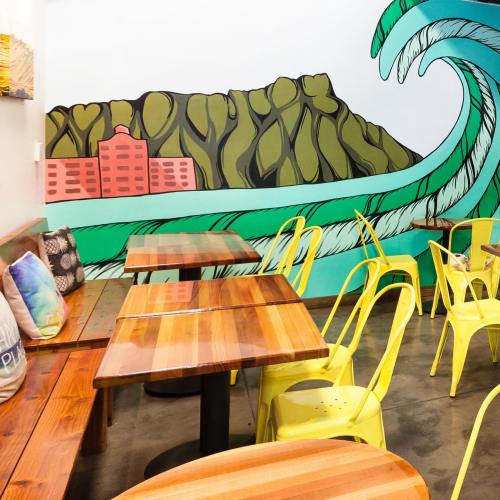 Colorful café interior featuring wooden tables, yellow chairs, and a vibrant mural of waves and hills on the wall.