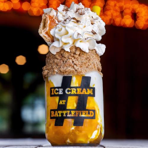 A tall dessert glass filled with ice cream, topped with whipped cream, a pastry, and cookie crumbles, against a blurred background.