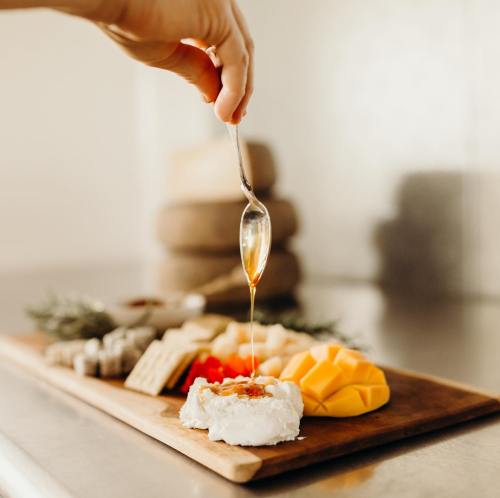 A hand drizzles honey over a cheese platter with fruits and crackers on a wooden board.