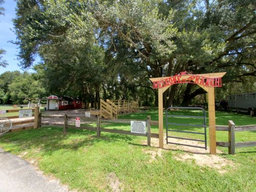 Entrance to a ranch with a wooden sign, surrounded by trees and a grassy area, featuring a fenced-off space.