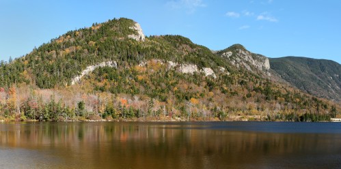 A serene lake reflects colorful autumn foliage and mountains under a clear blue sky.