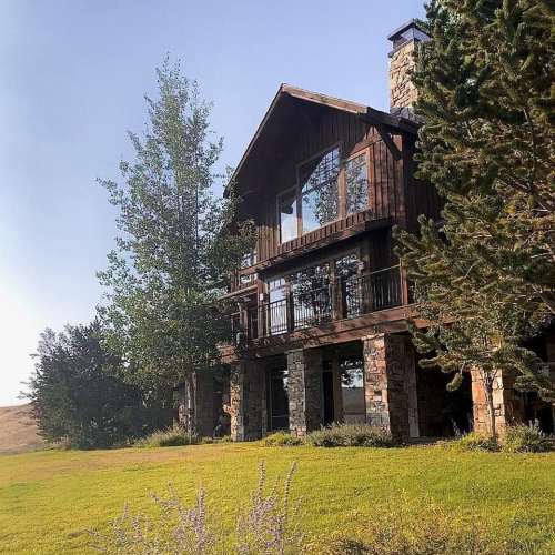A rustic wooden house with stone accents, surrounded by greenery and a clear blue sky.