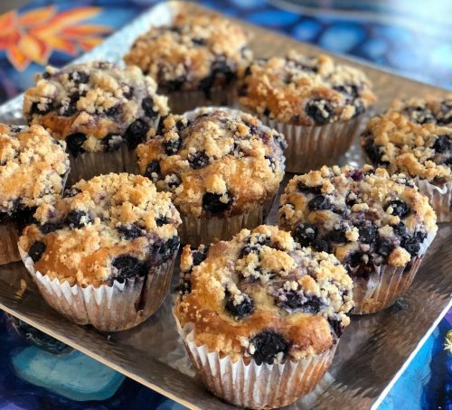 A tray of freshly baked blueberry muffins with a crumbly topping, set on a colorful table.
