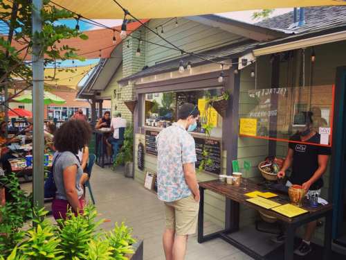 A busy outdoor café scene with people ordering food at a window and colorful canopies overhead.