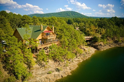 A scenic view of a wooden cabin on a rocky shore, surrounded by trees and overlooking a calm lake under a blue sky.
