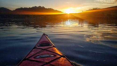 A kayak glides on calm water at sunset, with vibrant rays illuminating the landscape and mountains in the background.