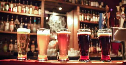 Five glasses of beer in varying colors lined up on a bar, with shelves of bottles in the background.