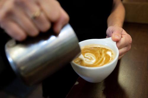 A person pours steamed milk into a cup of coffee, creating a swirl pattern on the surface.