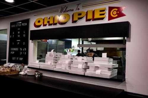 A pizza shop interior with a sign reading "Welcome to Ohio Pie" and stacked pizza boxes on the counter.