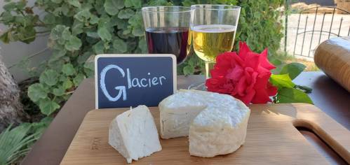 A cheese wheel on a wooden board with a slice cut out, two glasses of wine, and a rose nearby, labeled "Glacier."