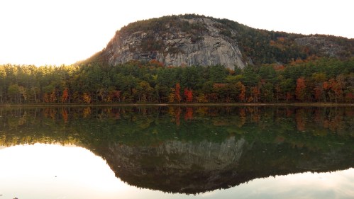 A serene lake reflects a rocky mountain and colorful autumn trees under a soft sunset glow.