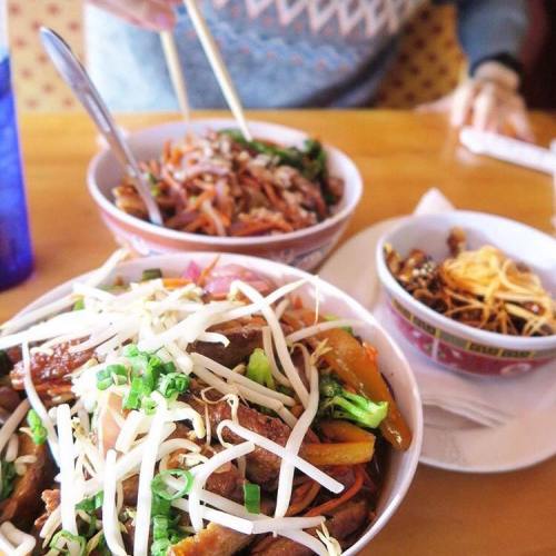 Two bowls of colorful Asian dishes with vegetables and noodles, accompanied by chopsticks and a drink.