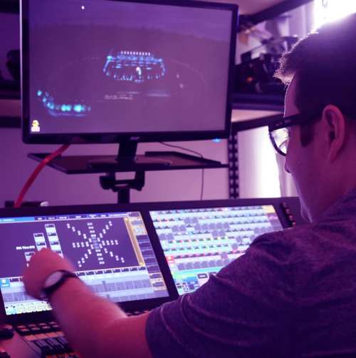 A person operates a lighting control board, with a monitor displaying a stage setup in the background.