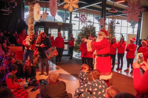 A festive scene with Santa interacting with children, surrounded by holiday decorations and a cheerful crowd.