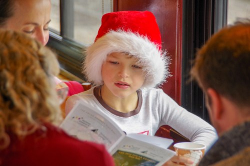 A child in a Santa hat reads a booklet at a table with adults, enjoying a cozy indoor setting.