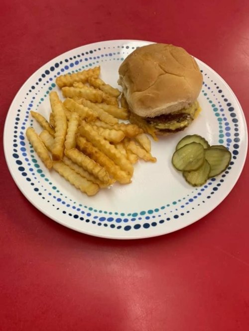 A plate with a cheeseburger, crinkle-cut fries, and two pickle slices on a red table.