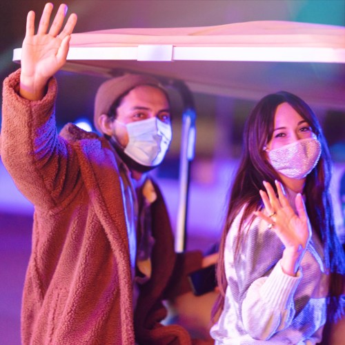 Two people wave while sitting under a canopy, both wearing masks, with colorful lights in the background.
