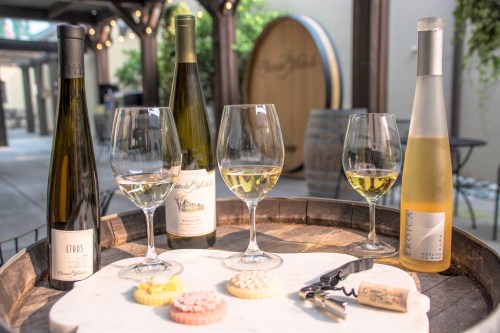 A wooden tray with wine glasses, bottles of wine, and colorful cookies, set in a cozy outdoor space.