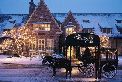 A horse-drawn carriage in front of The American Club, adorned with lights and surrounded by snow at dusk.