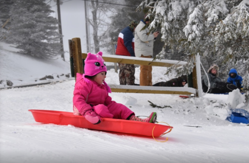 A young child in a pink snowsuit and hat sleds down a snowy hill, with people enjoying the winter scene in the background.