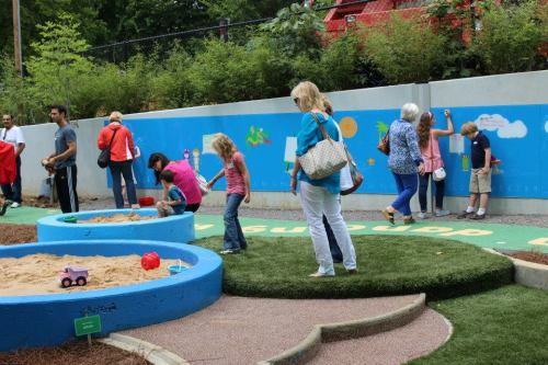 A playground scene with children playing in sandboxes and adults interacting, surrounded by colorful murals and greenery.