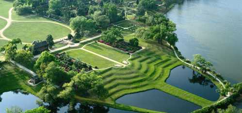 Aerial view of a landscaped park with gardens, pathways, and a lake, featuring lush greenery and patterned lawns.