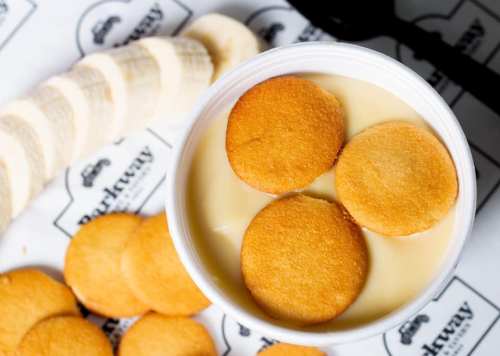 A cup of creamy dessert topped with round cookies, alongside sliced bananas and additional cookies on a patterned surface.
