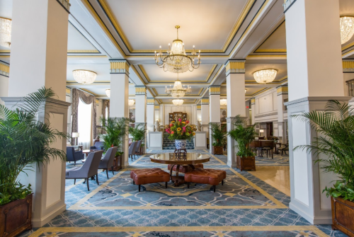 Elegant hotel lobby featuring chandeliers, plush seating, and decorative plants, with a central table and patterned carpet.