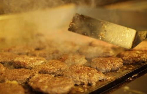 A close-up of sizzling hamburger patties on a grill, with steam rising and a spatula flipping one of the burgers.