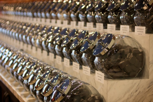A row of glass jars filled with various candies, labeled and displayed on a marble counter.