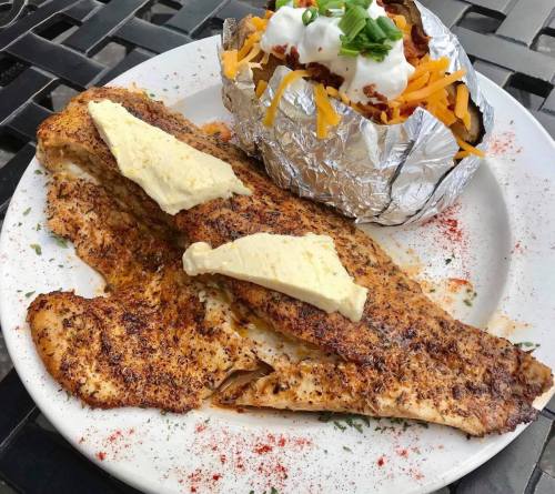 A plate with seasoned fish fillets topped with butter, alongside a baked potato with sour cream, cheese, and green onions.