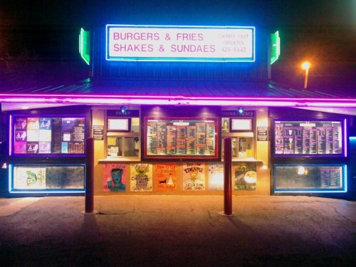 A brightly lit burger and fries stand with a colorful menu and neon signs, showcasing shakes and sundaes.