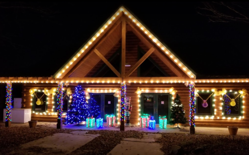 A cozy log cabin decorated with colorful holiday lights and Christmas trees at night.