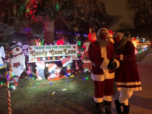 A couple dressed as Santa stands in front of a festive Candy Cane Lane sign, surrounded by colorful holiday decorations.