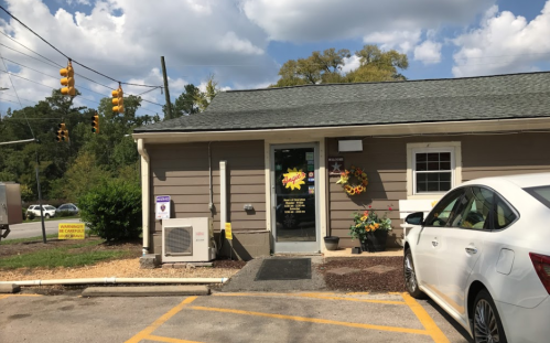 A small, single-story building with a sign, flowers, and a parked white car, under a partly cloudy sky.